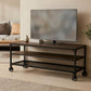 Mesa para TV industrial modelo Nashville en acabado de madera color nogal con estructura de metal negro, repisa de rejilla y ruedas, con televisión sobre la cubierta en una sala con sofá.