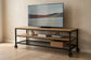 Vista frontal de la mesa para TV industrial modelo Nashville con dos entrepaños de maderacolor roble, repisa inferior de rejilla metálica con canasta de mimbre y ruedas de metal negro.